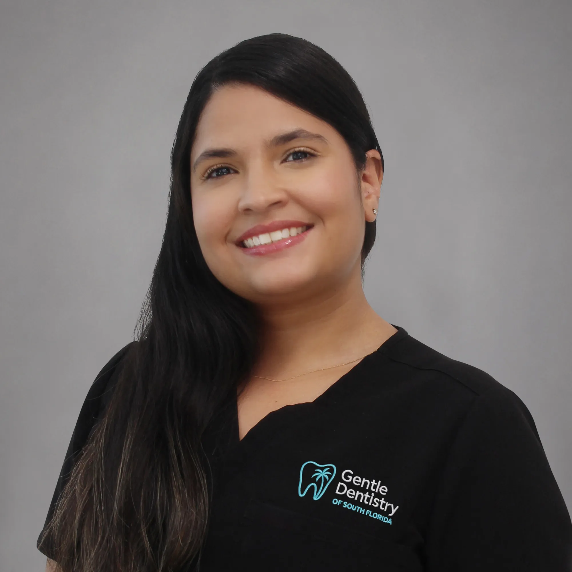 Portrait of a smiling female team member with long dark hair parted on the side wearing black Gentle Dentistry scrubs