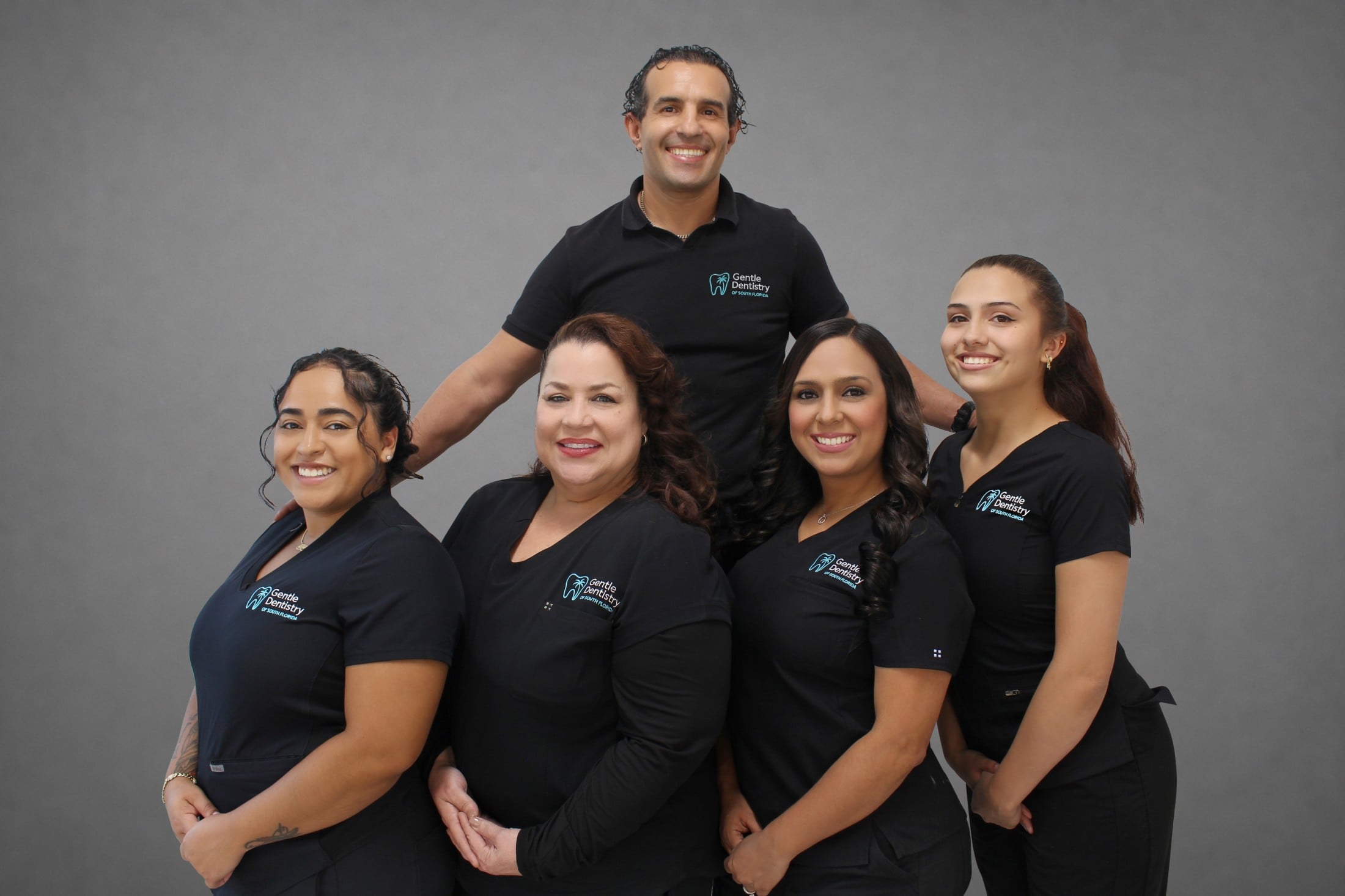 Five dental staff members in matching black Gentle Dentistry polo shirts and scrubs posing together against a grey backdrop