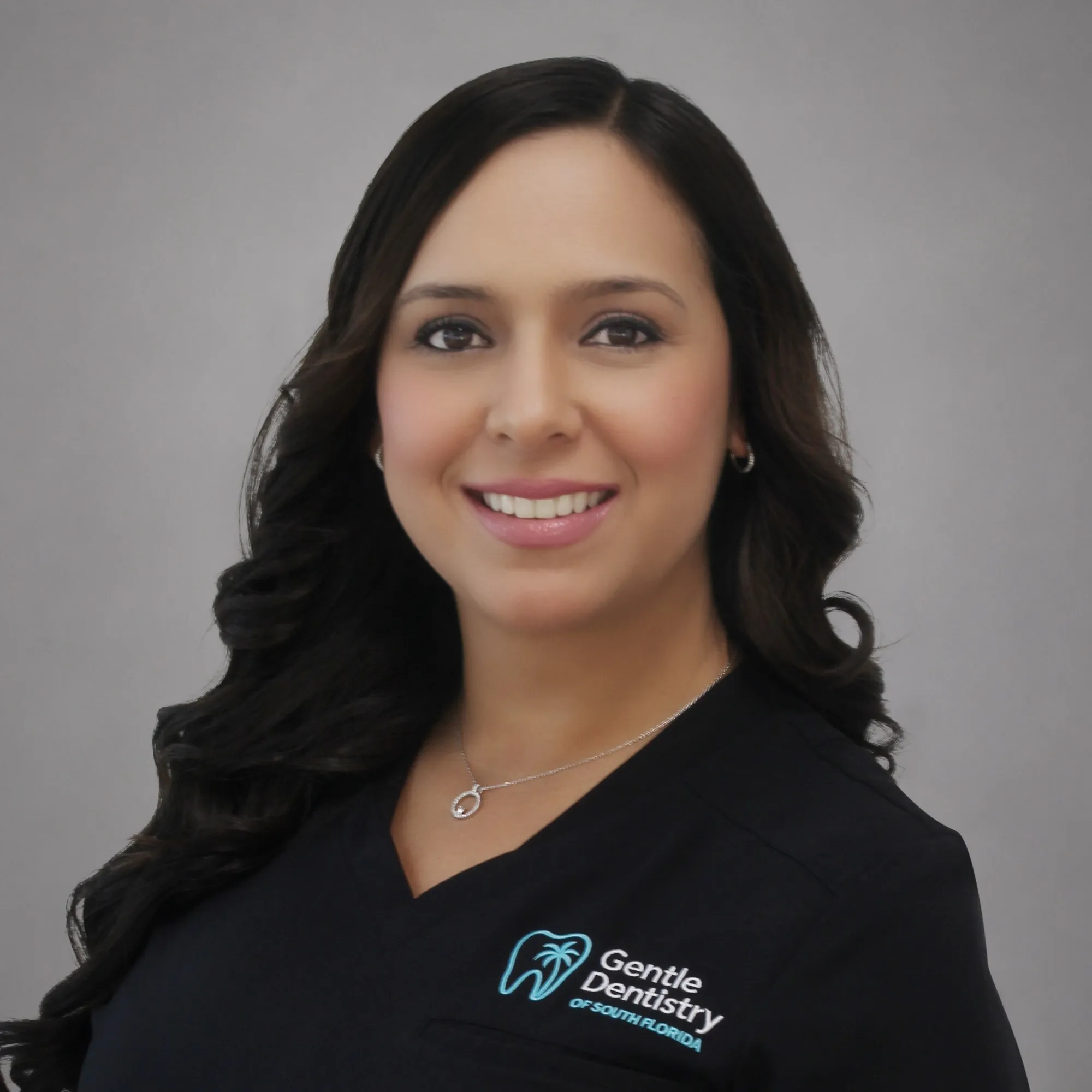 Headshot of a smiling female healthcare professional with long, dark, wavy hair wearing a black Gentle Dentistry scrub top against a grey background